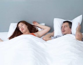 Closeup of sleeping and snoring woman and her husband who is lying beside her in bed and covering ears with pillow
